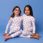 2 models sitting side-by-side in long-sleeved, GOTS certified, 100% organic cotton pajamas against a bright blue background. 
 Pajamas are white, lightweight fabric with a vibrant, colorful, dancing unicorns print.
good morning monsters pajamas, loungewear, sleepwear, pajamas, boys, girls, baby, unisex