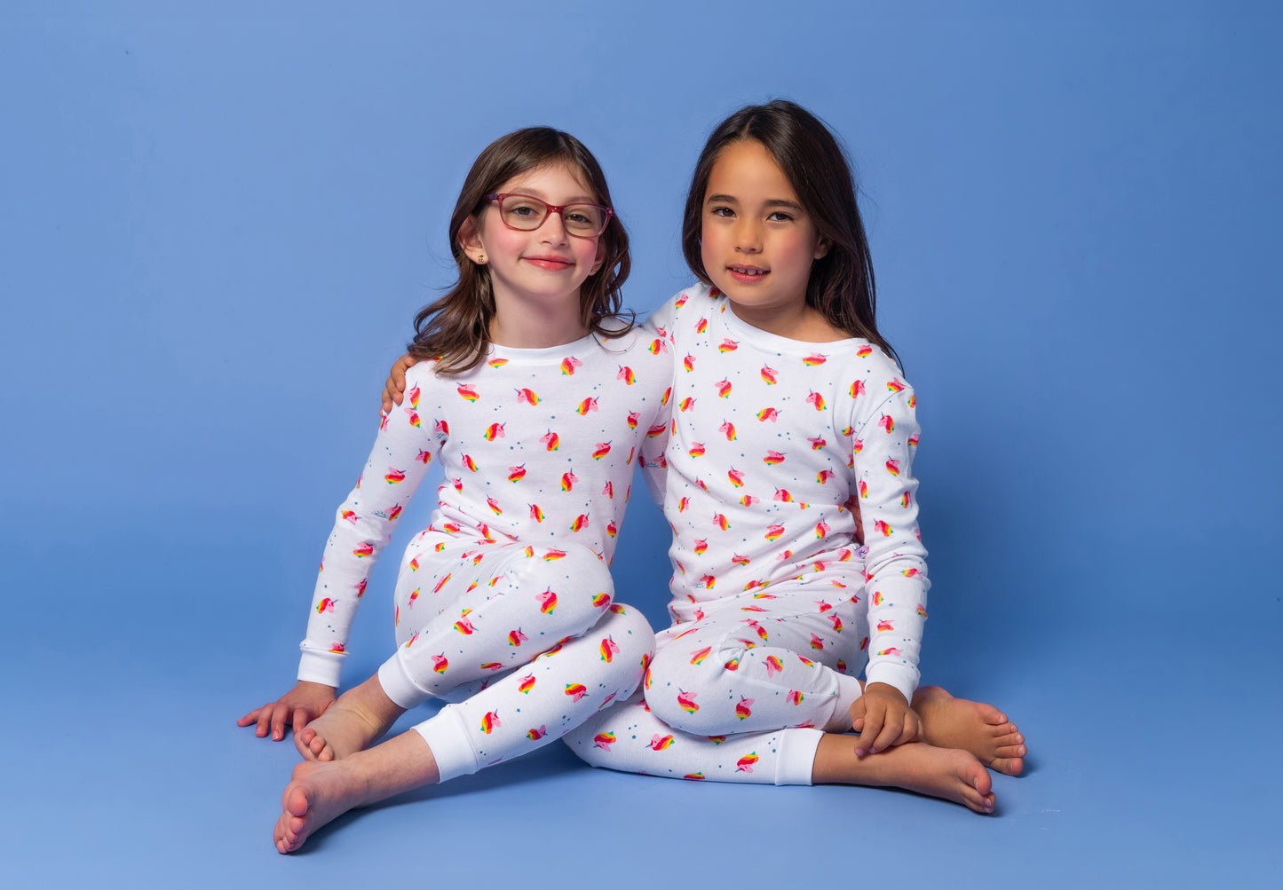 2 models sitting side-by-side in long-sleeved, GOTS certified, 100% organic cotton pajamas against a bright blue background. 
 Pajamas are white, lightweight fabric with a vibrant, colorful, dancing unicorns print.
good morning monsters pajamas, loungewear, sleepwear, pajamas, boys, girls, baby, unisex
