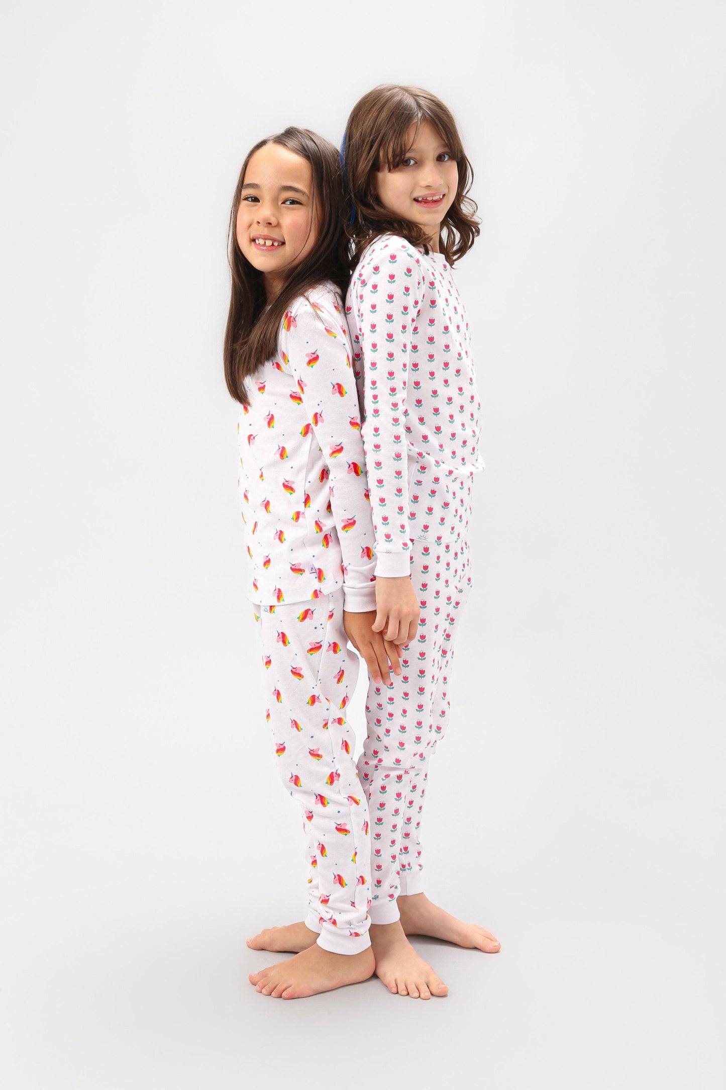 2 models standing back to back (one model wearing unicorn print PJ set, other model wearing tulips print PJ set).  
Pajamas are white fabric with vibrant, colorful prints.  Pajamas are long-sleeved and made from GOTS certified, 100% organic cotton.
good morning monsters pajamas, loungewear, sleepwear, pajamas, boys, girls, baby, unisex