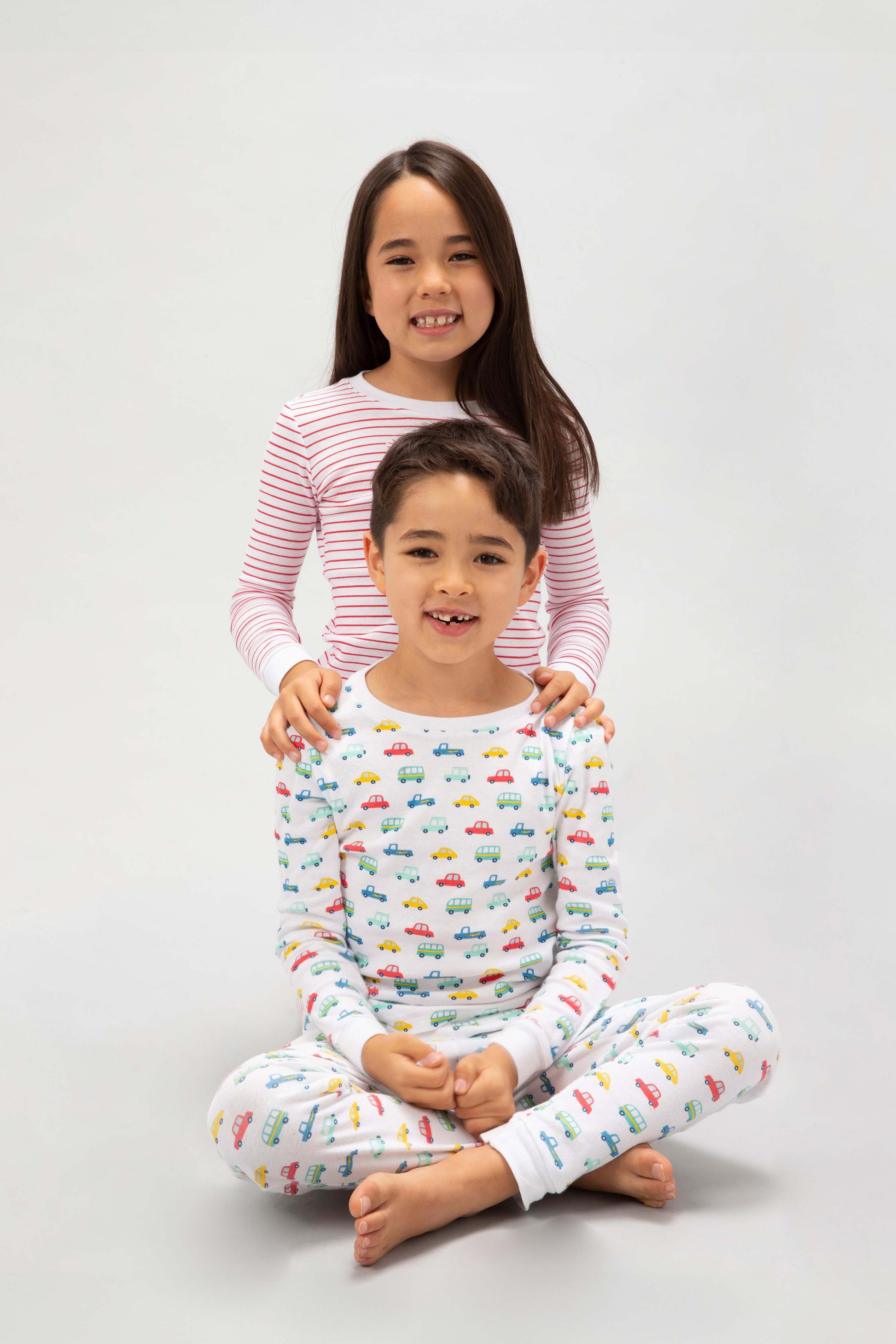 2 models sitting, one in a brightly colorful vehicles print, the other in red stripes prints.
Pajamas are white, lightweight, fabric with vibrant, colorful prints.  Pajamas are long-sleeved and made from  GOTS certified, 100% organic cotton.
good morning monsters pajamas, loungewear, sleepwear, pajamas, boys, girls, baby, unisex