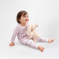 Toddler posing in long-sleeved, GOTS certified, 100% organic cotton pajamas - and she is holding a bunny doll. 
 Pajamas are white, lightweight fabric with a vibrant tulips print (bright pink tulips with turquoise leaves).