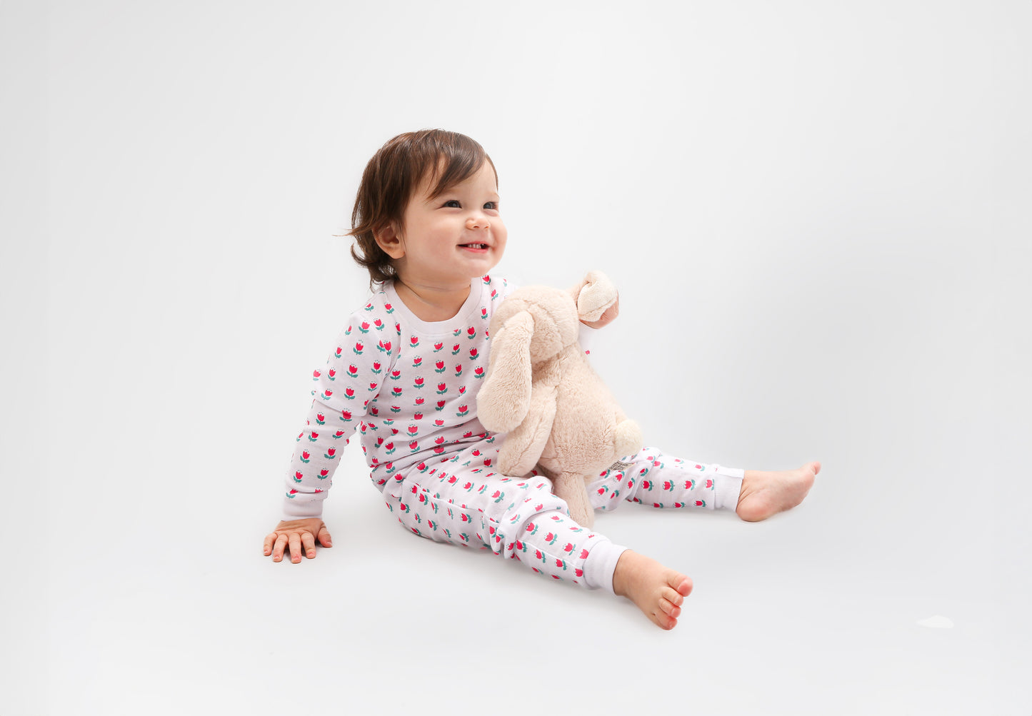 Toddler posing in long-sleeved, GOTS certified, 100% organic cotton pajamas - and she is holding a bunny doll. 
 Pajamas are white, lightweight fabric with a vibrant tulips print (bright pink tulips with turquoise leaves).