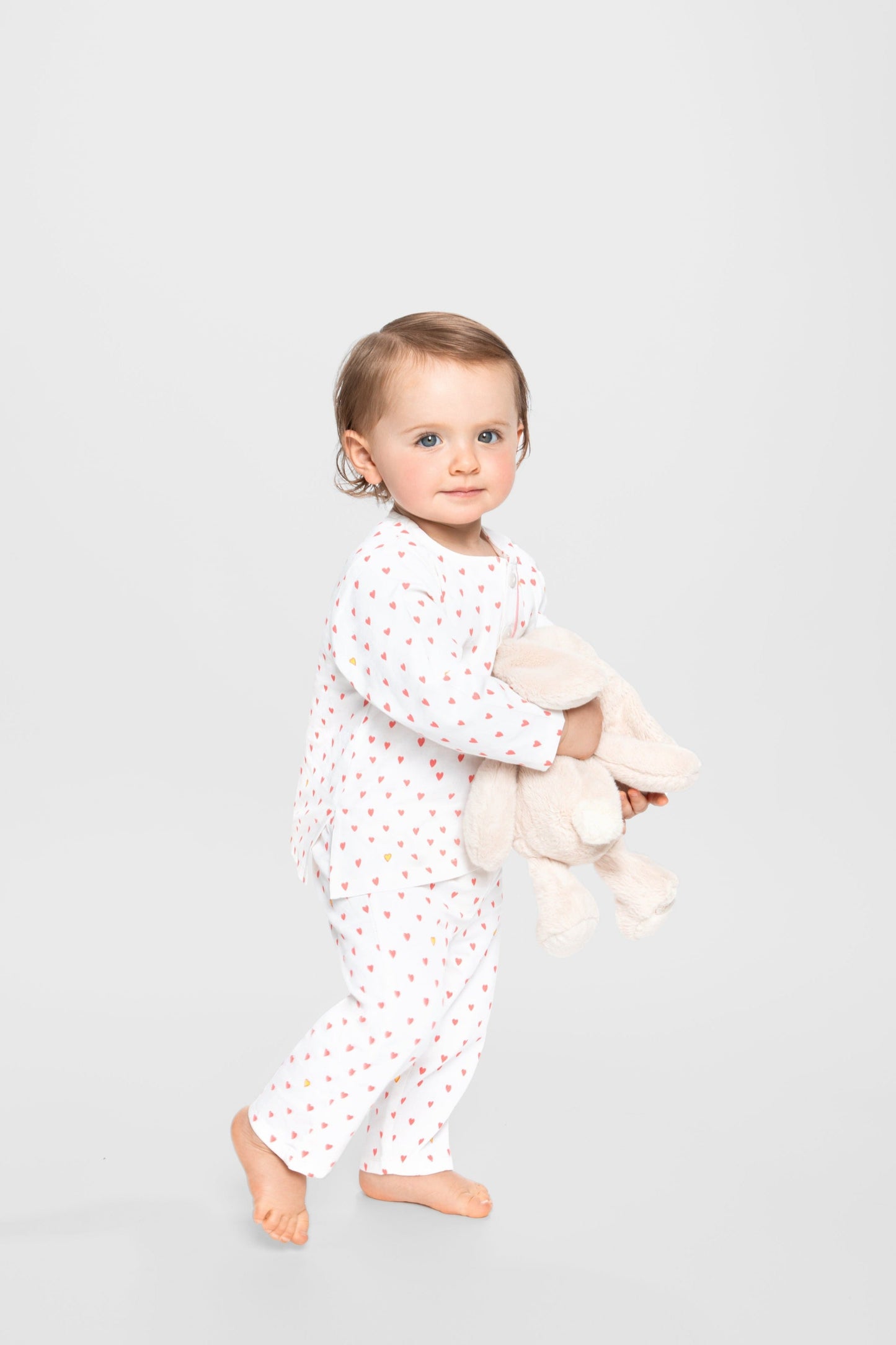 model wearing hearts print pajamas (solid white fabric with pink hearts) holding jellycat doll; good morning monsters woven pajamas, loungewear, sleepwear, loose-fitting pajamas, boys, girls, baby, unisex