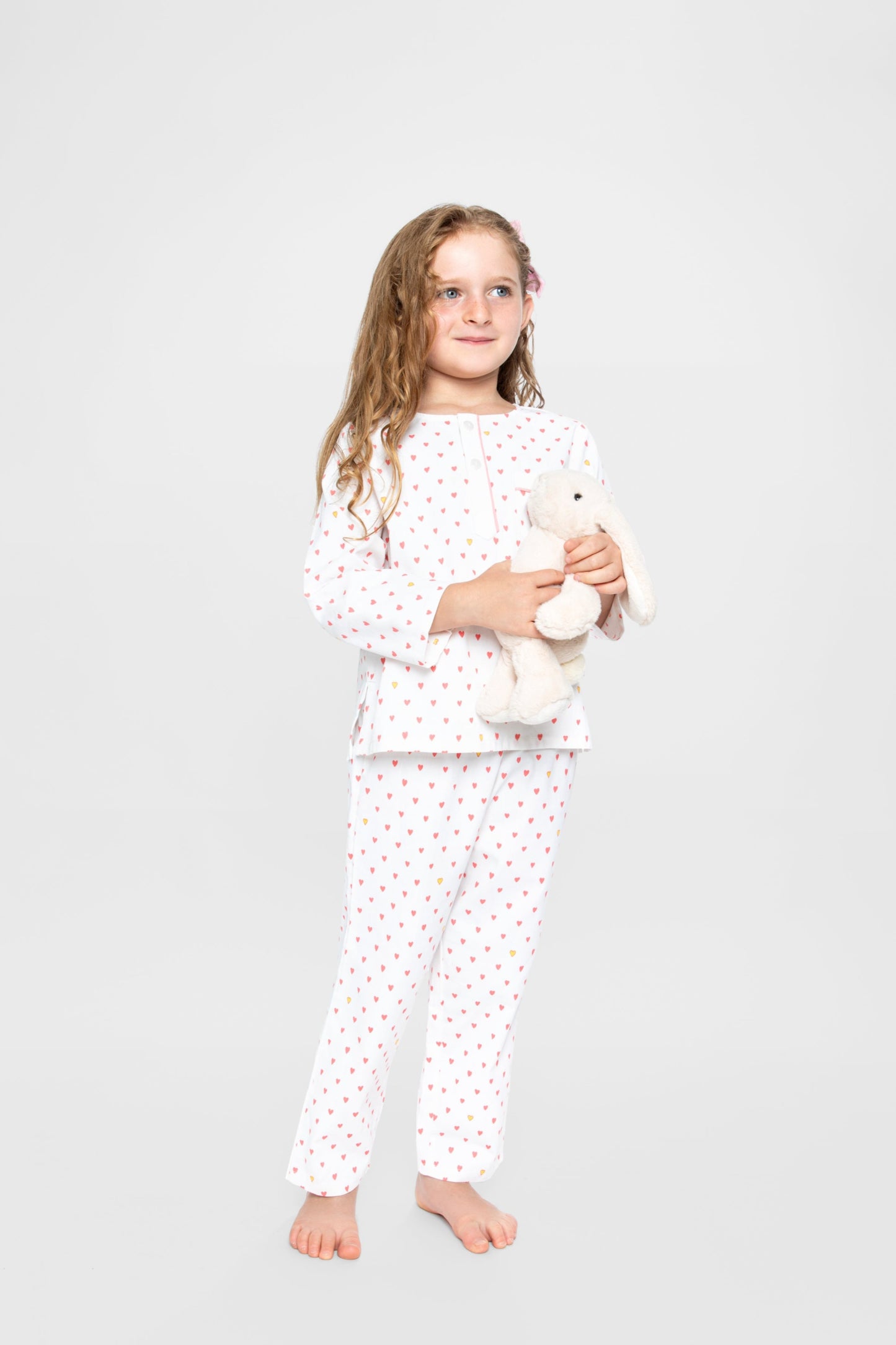 model wearing hearts print pajamas (solid white fabric with pink hearts) holding jellycat doll; good morning monsters woven pajamas, loungewear, sleepwear, loose-fitting pajamas, boys, girls, baby, unisex