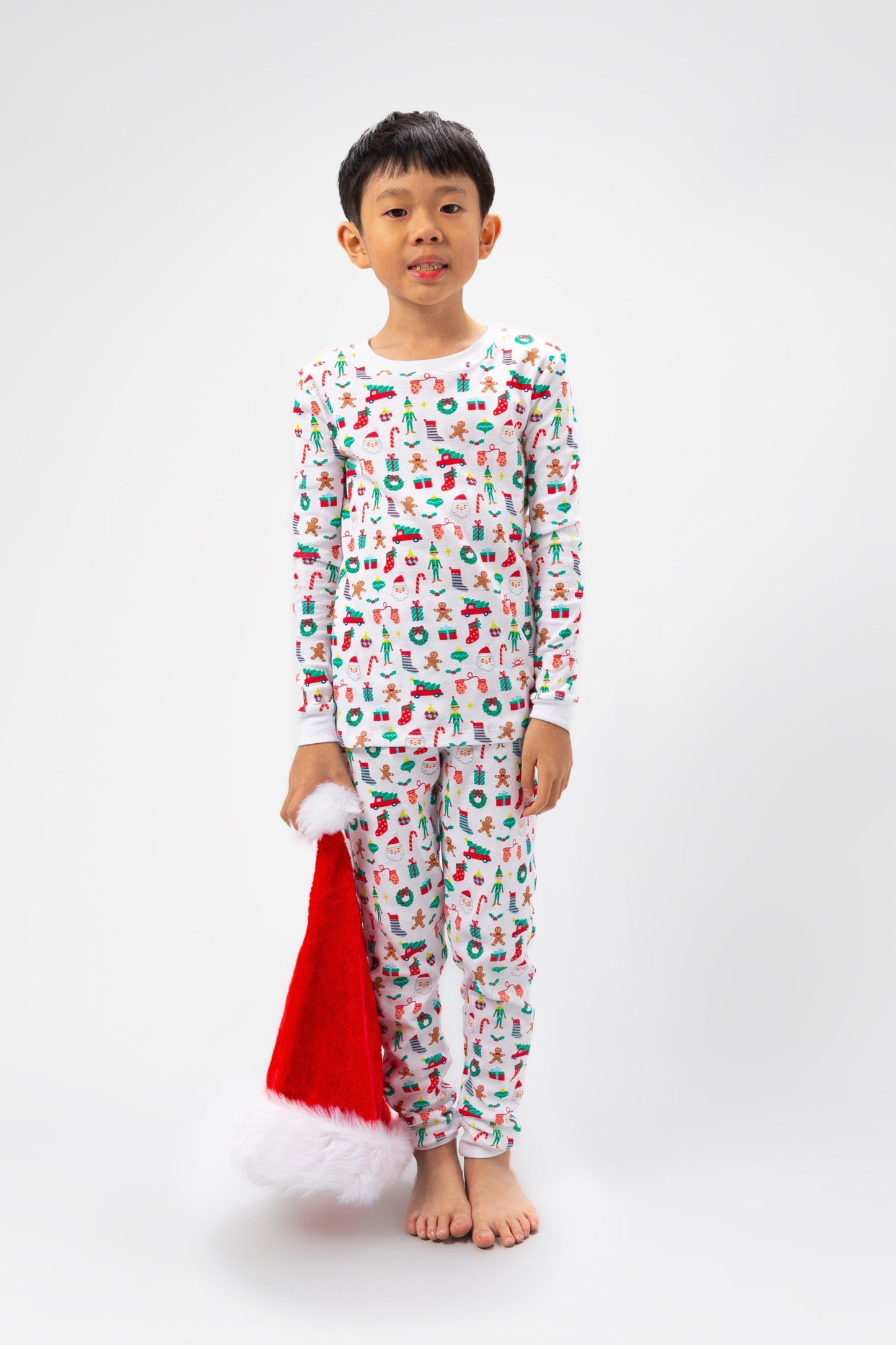 Front view of model with Santa hat, wearing Christmas, festive, Santa, Elves, Classic Christmas, Holiday, Ornaments, Stockings, Gingerbread, Red Trucks pajama set.  
Pajamas are white, lightweight fabric with a vibrant Christmas print.   Pajamas are long-sleeved and made from GOTS certified, 100% organic cotton.
good morning monsters pajamas, loungewear, sleepwear, pajamas, boys, girls, baby, unisex