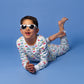 Model posing in white sunglasses in long-sleeved, GOTS certified, 100% organic cotton pajamas against a bright blue background. 
 Pajamas are white, lightweight fabric with a vibrant, colorful, vehicles print.
good morning monsters pajamas, loungewear, sleepwear, pajamas, boys, girls, baby, unisex