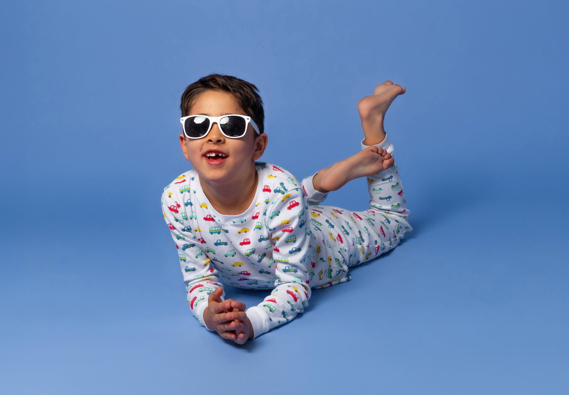 Model posing in white sunglasses in long-sleeved, GOTS certified, 100% organic cotton pajamas against a bright blue background. 
 Pajamas are white, lightweight fabric with a vibrant, colorful, vehicles print.
good morning monsters pajamas, loungewear, sleepwear, pajamas, boys, girls, baby, unisex