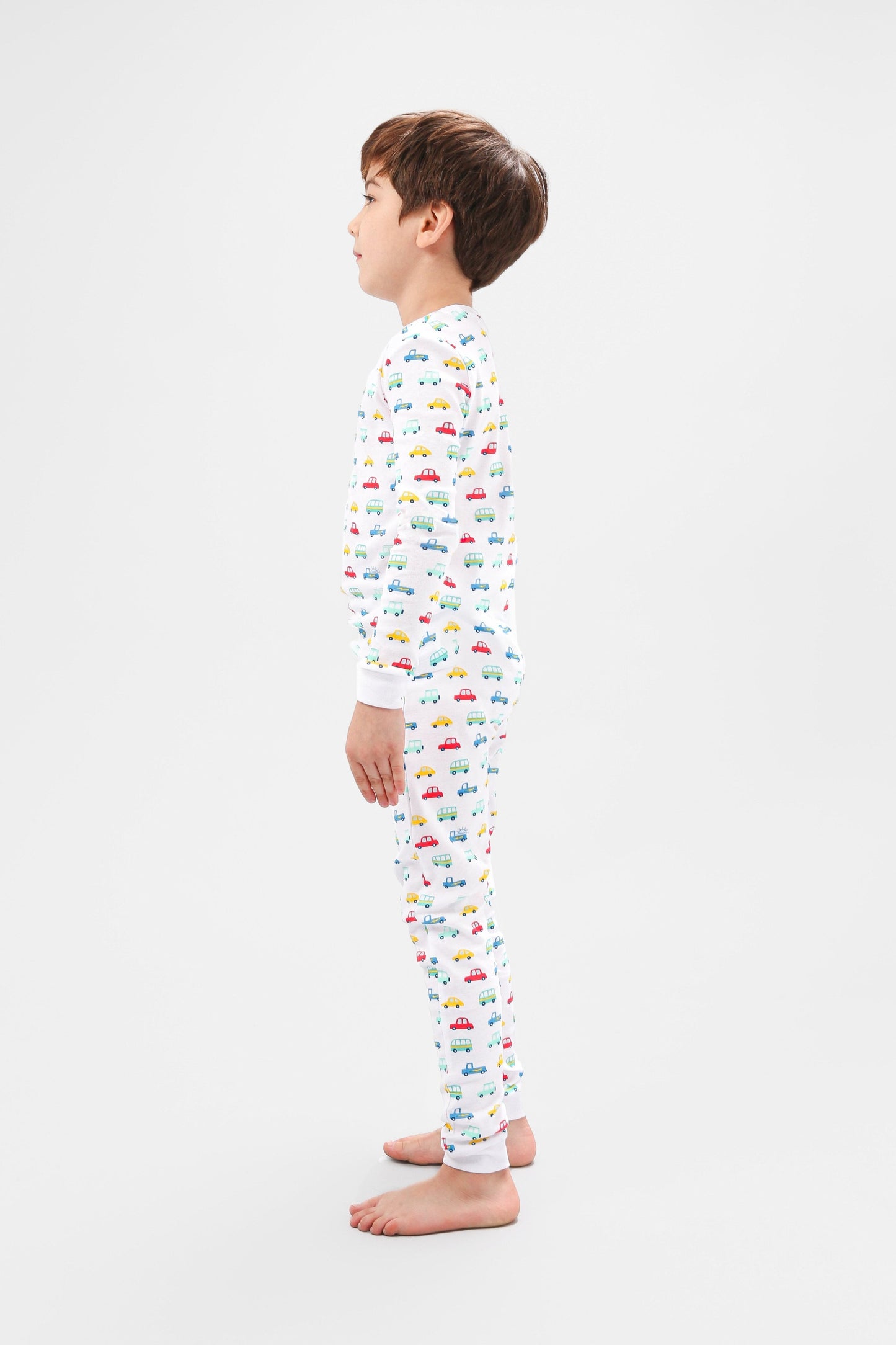 Side view of model wearing vehicles pajama set.  
Pajamas are white, lightweight fabric with a vibrant, colorful vehicles print with cars, jeeps, trucks, and buses.  Pajamas are long-sleeved and made from 100% GOTS certified organic cotton.
good morning monsters pajamas, loungewear, sleepwear, pajamas, boys, girls, baby, unisex