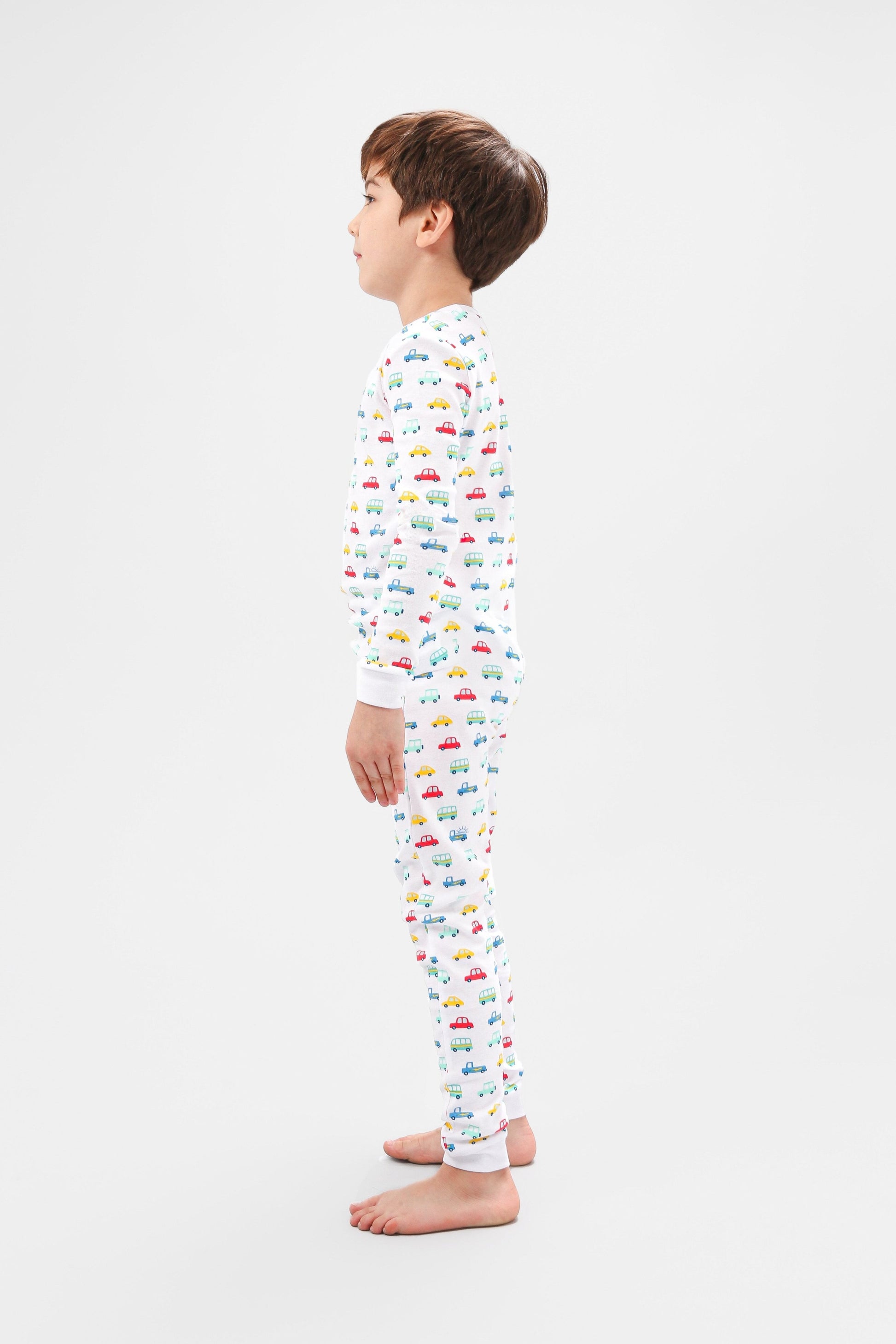 Side view of model wearing vehicles pajama set.  
Pajamas are white, lightweight fabric with a vibrant, colorful vehicles print with cars, jeeps, trucks, and buses.  Pajamas are long-sleeved and made from 100% GOTS certified organic cotton.
good morning monsters pajamas, loungewear, sleepwear, pajamas, boys, girls, baby, unisex